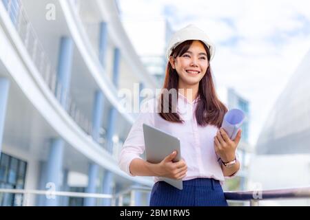 Felice giovane attraente ingegnere asiatico donna che indossa l'elmetto che tiene il suo computer tablet e blueprint mentre si trova fuori di ufficio edificio, autobus Foto Stock