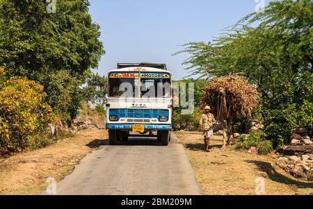 Un operaio agricolo e il suo cammello che porta una cauzione di paglia con l'autobus locale su una strada rurale in Rajasthan, India Foto Stock