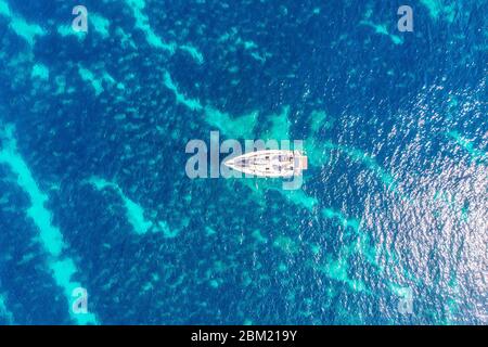 Pleasure yacht bianco con vela si erge sulla barriera corallina in acque turchesi blu trasparenti. Viaggio concettuale. Vista dall'alto dell'antenna. Foto Stock