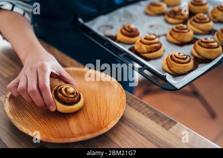 Primo piano di una mano di una donna che raccoglie i rotoli di cannella appena sfornati in casa su un foglio di biscotto. Foto Stock