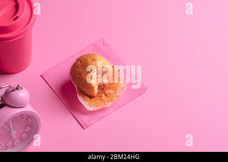 Tavolo per la colazione con muffin, una tazza di caffè e sveglia. Morso il muffin su sfondo rosa. Tazza di caffè rosa e orologio. Foto Stock