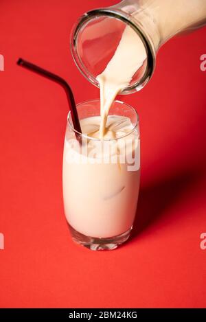 Versando l'orchata, la mandorla messicana e il latte di riso, in un bicchiere con cubetti di ghiaccio, su fondo rosso. Serve un cocktail spagnolo freddo senza alcol. Foto Stock