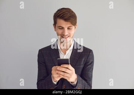 Giovane uomo in giacca con un cellulare in mano a scrivere sms sorride ridendo gioioso felice positivo su uno sfondo grigio. Imprenditore uomo d'affari freela Foto Stock