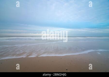 Una spiaggia nei Paesi Bassi alla luce della sera. Il cielo nuvoloso si riflette nel mare, le onde corrono verso la riva. Foto Stock