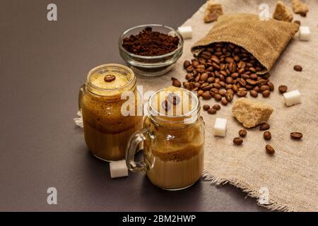 Caffè Dalgona in vasetto di muratori. Bevanda a base di caffè frusta coreana. Bevanda alla moda in quarantena, zucchero e chicchi di caffè su fondo in cemento di pietra nera Foto Stock