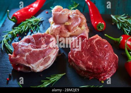 Tre tipi di carne di pollo, maiale e manzo su pietra nera con erbe e pepe Foto Stock