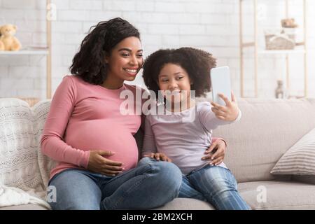 Piccola ragazza africana che prende Selfie con la mamma incinta felice a casa Foto Stock