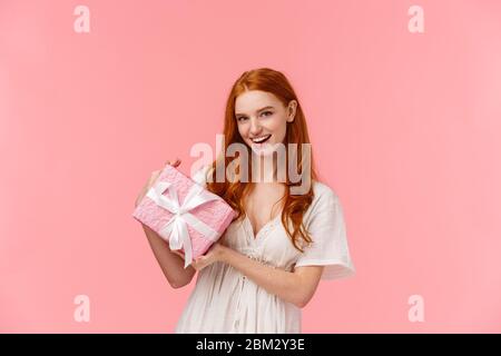 Donna rossa dall'aspetto accattivante e vivace, con un abito bianco elegante, che tiene avvolto il presente, chiedendo a indovinare cosa c'è dentro, sorridendo cheeky, dando romantico Foto Stock