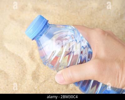 Mano donna che tiene una bottiglia di plastica blu con acqua potabile su una sabbia di mare. Concetto di sete, estate calda, vacanza al mare e vacanza in spiaggia. Foto Stock