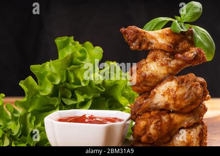 Ali di pollo fritte su un tavolo da cucina in legno con foglie di basilico e lattuga su un tavolo di legno, su sfondo nero. Fast food. Foto Stock