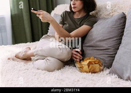 Una donna bellissima passa canali e film su un televisore utilizzando un telecomando. Beve birra e mangia patatine a casa. Rilassati a casa guardando la TV e i film. Foto Stock