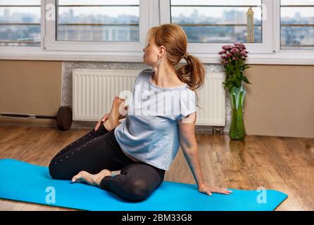 Una bella donna di mezza età di aspetto europeo pratica yoga su un karemat blu nel suo appartamento vicino alla finestra. Foto Stock