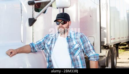 Pilota americano dall'aspetto deciso che indossa una camicia a quadri blu e un cappello da baseball nero con la bandiera americana accanto al grande carro. Foto Stock