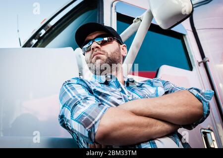 Sicuro caucsiano maschio americano camion accanto al suo grande carro. Concetto di autotrasporti proprietari operatori con uomo in maglia a quadri e cappello da baseball.