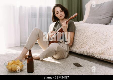 Una giovane donna gioca ukulele, beve birra e mangia patatine, seduto a casa. Il concetto di riposo durante l'autoisolamento e la quarantena. Foto Stock