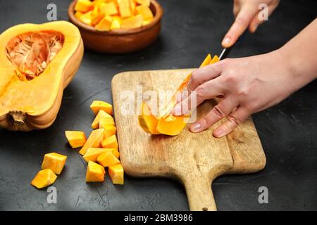 Donna taglia una zucca su un tagliere di legno sul tavolo di pietra. Mani femminili con coltello in acciaio, pezzi di zucca in tovaglietta di legno su backgroun nero Foto Stock