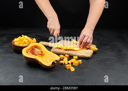 Donna taglia una zucca su un tagliere di legno sul tavolo di pietra. Mani femminili con coltello in acciaio, pezzi di zucca in tovaglietta di legno su backgroun nero Foto Stock