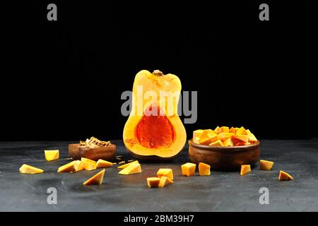 Metà zucca, pezzi freschi in ciotola di legno e sparsi su sfondo nero. Squash di butternut crudo tritato, fuoco selettivo. Piatto da cucina vegetariano Foto Stock