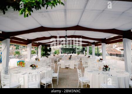 Ricevimento con tavolo per cena per matrimoni. Tavoli rotondi con tovaglie bianche e sedie bianche Chiavary sotto una grande tenda bianca, sullo sfondo di un Foto Stock