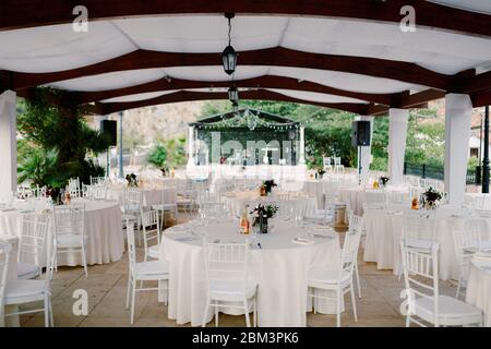 Ricevimento con tavolo per cena per matrimoni. Tavoli rotondi con tovaglie bianche e sedie bianche Chiavary sotto una grande tenda bianca, sullo sfondo di un Foto Stock