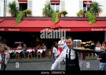 Posti a sedere all'aperto della brasserie ristorante di Au Cadet de Gascogne con i clienti e un cameriere che porta cibo in un grande vassoio.Montmartre.Paris.France Foto Stock