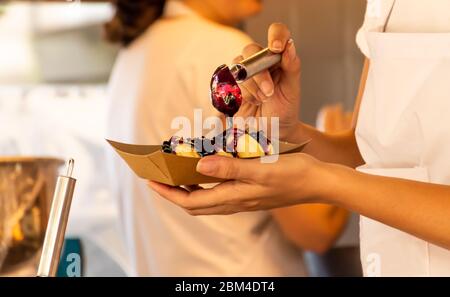 Donna che versa la marmellata di frutta dolce sopra le ciambelle durante il festival di fast food. Camion per alimenti. Catering esterno. Cibo a buffet Catering Dining Party Sharing Foto Stock