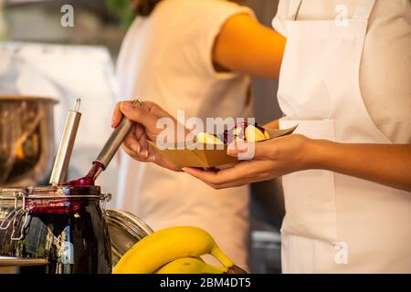 Donna che versa la marmellata di frutta dolce sopra le ciambelle durante il festival di fast food. Camion per alimenti. Catering esterno. Cibo a buffet Catering Dining Party Sharing Foto Stock