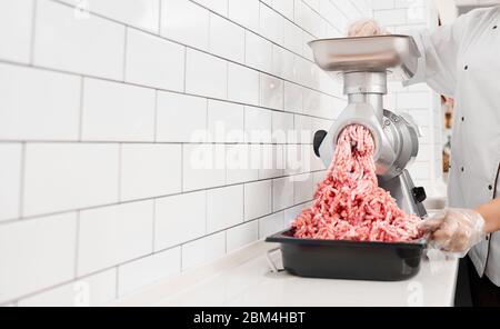 Raccolto di donna incognito con capelli ricci mettendo piccoli pezzi nel macinacaffè e li macina in macinacaffè. Vista della lavorazione femminile in macelleria, preparazione di carne per la vendita in negozio. Foto Stock