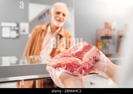 Fuoco selettivo di pezzo fresco crudo rosso di carne nelle mani di giovane donna incognito. Dall'alto, la ragazza si trova dietro il bancone con una grande varietà di carne e mostra maiale all'uomo anziano. Foto Stock