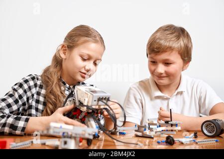 Vista frontale di ragazzo e ragazza che si divertono, creando robot. Ingegneria scientifica. Amici interessati sorridenti, chiacchierando e lavorando insieme al progetto utilizzando un interessante kit di costruzione per bambini sul tavolo. Foto Stock