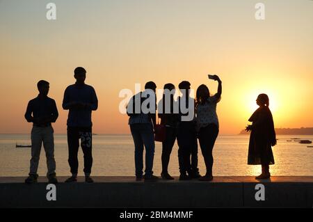 Un gruppo di giovani al tramonto che si erge sulla parete di argine di Marine Drive, vicino al Mar Arabico, Mumbai, India, prendendo selfie Foto Stock