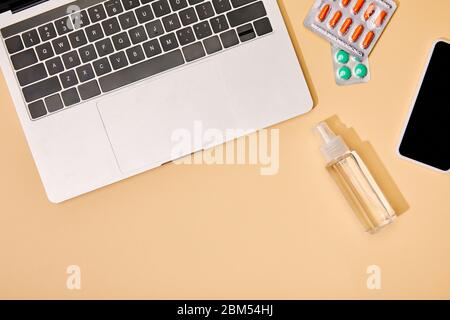 vista dall'alto di laptop e smartphone con schermo vuoto vicino alle pillole e bottiglia con igienizzante per le mani su beige Foto Stock