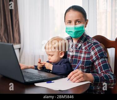 Autoisolamento per proteggere la vostra famiglia dal virus covid-19. La madre che indossa una maschera rimane a casa lavorando al computer tenendo il suo piccolo figlio carino sui suoi giri. Concetto di soggiorno a casa Foto Stock