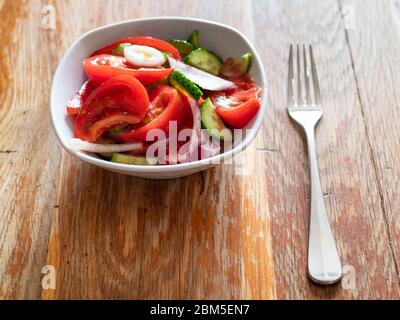 porzione di insalata fatta in casa a partire da verdure fresche (pomodori a fette e oliati, cetrioli, cipolla) in una ciotola bianca su un tavolo di legno shabby in cucina Foto Stock