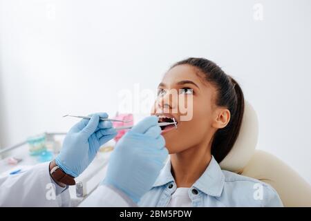 dentista in guanti di lattice che tiene l'attrezzatura dentale vicino a paziente afro-americano con bocca aperta e bretelle Foto Stock