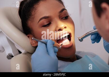 fuoco selettivo del dentista in guanti di lattice che tengono gli strumenti dentali vicino alla donna afro-americana attraente in bretelle Foto Stock