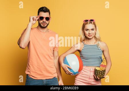 bella, ragazza bionda che tiene palla gonfiabile e ananas con cocktail vicino uomo bello che tocca occhiali da sole su sfondo giallo Foto Stock