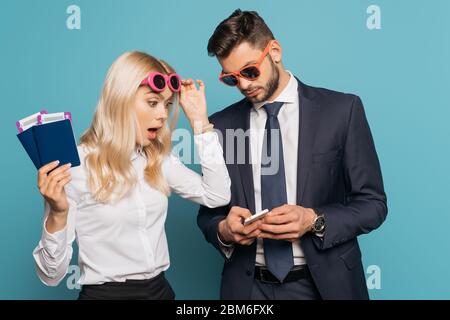 donna d'affari scioccata che tiene documenti e occhiali da sole toccanti mentre guarda lo smartphone in mano a un uomo d'affari su sfondo blu Foto Stock