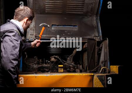 Un meccanico arrabbiato maschio sta riparando il motore di un vecchio camion con un cofano aperto che lo colpisce con un martello. Foto Stock