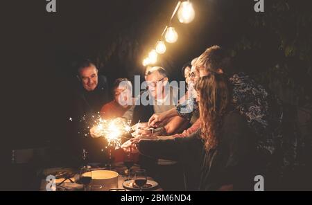 Buona famiglia divertirsi a cena festa all'aperto - Gruppo di persone diverse età festeggiando insieme con fuochi d'artificio scintillio all'esterno Foto Stock