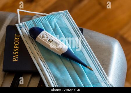 Primo piano di una maschera medica facciale, passaporto e termometro sulla valigia per viaggi di lavoro o vacanze durante l'epidemia di coronavirus Foto Stock