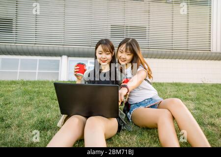 Due simpatiche e gioiose giovani amiche asiatiche che si divertono mentre si siedono sul verde erba all'aperto in città e guardare sullo schermo del computer portatile per guardare Foto Stock