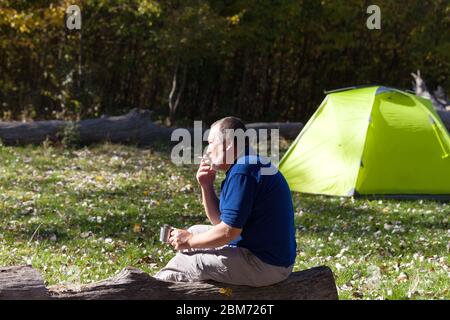 Uomo seduto su legno di legno con tazza di tè e sigaretta. L'escursionista si crogiola al sole, beve tè e fuma sigarette. Luce solare verde campeggio tenda e autu Foto Stock