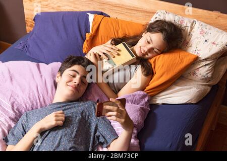 Una giovane coppia è sdraiata a letto. Entrambi guardano il telefono mentre si divertono Foto Stock