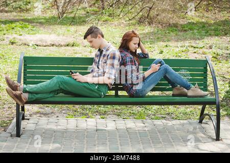 Una giovane coppia seduta in un parco su una panchina guardando ciascuno nel loro telefono cellulare e guardando lontano l'uno dall'altro Foto Stock