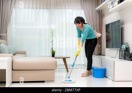 Attraente giovane donna asiatica che pulisce il pavimento delle mattonelle al soggiorno mentre fa la pulizia a casa durante il soggiorno a casa usando il tempo libero circa la loro h quotidiana Foto Stock
