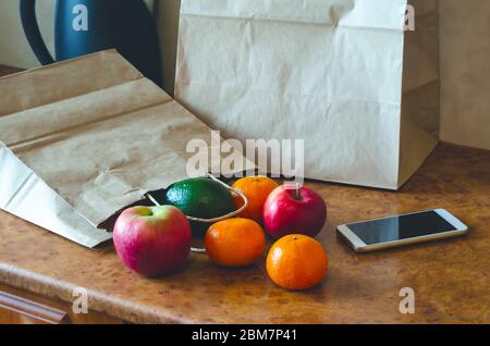 Sacchetti di carta, verdure fresche e frutta accanto a un telefono cellulare sul tavolo con utensili da cucina. Acquisto online e consegna senza contatto. Foto Stock