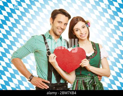 Coppia a dirndl e pantaloni in pelle tiene un cuore rosso davanti a una bandiera bavarese Foto Stock