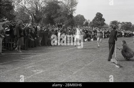 Scuola secondaria sport, Inghilterra, c1960, fuori su un percorso di erba, allietato da una grande folla di compagni di scuola e insegnanti, due giovani uomini che corrono duro per il nastro finale alla fine di una corsa di relè. Foto Stock