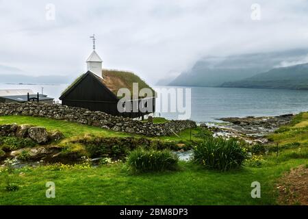 Vista estiva della tradizionale chiesa in cima a un manto erboso nel villaggio di faroese. Paesaggio di bellezza con fiordo foggy e alte montagne. Isola di Streymoy, Isole Faroe, Danimarca. Foto Stock